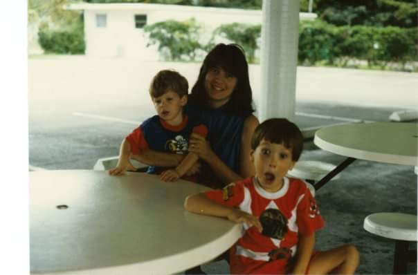 A very young Richard (in power ranger outfit) with baby brother, Wesley, and mom, Terri. They are sitting at an outdoor diner table.